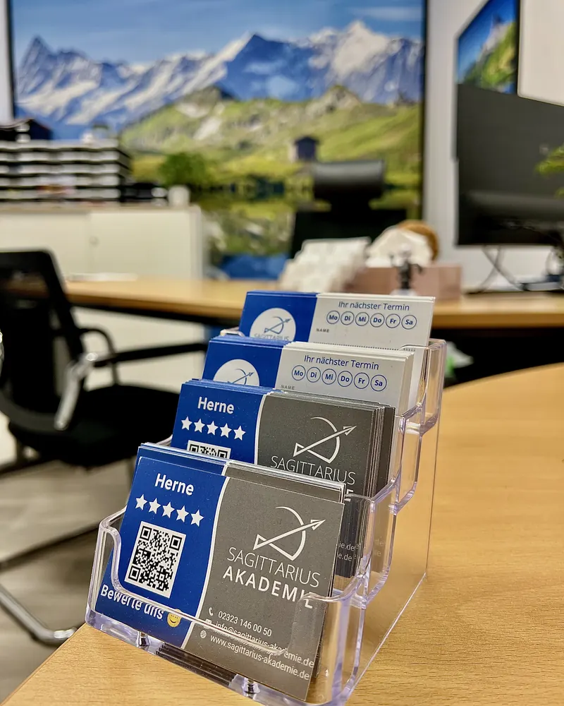 Terminkarten „Ihr nächster Termin“ und Karten zum Bewerten mit QR-Code in einem Aufsteller auf dem Schreibtisch, dahinter ein Büro mit Bergbild